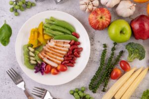 A top-down view of a plate with sliced grilled chicken, green beans, kidney beans, kiwi, and squash, surrounded by produce.